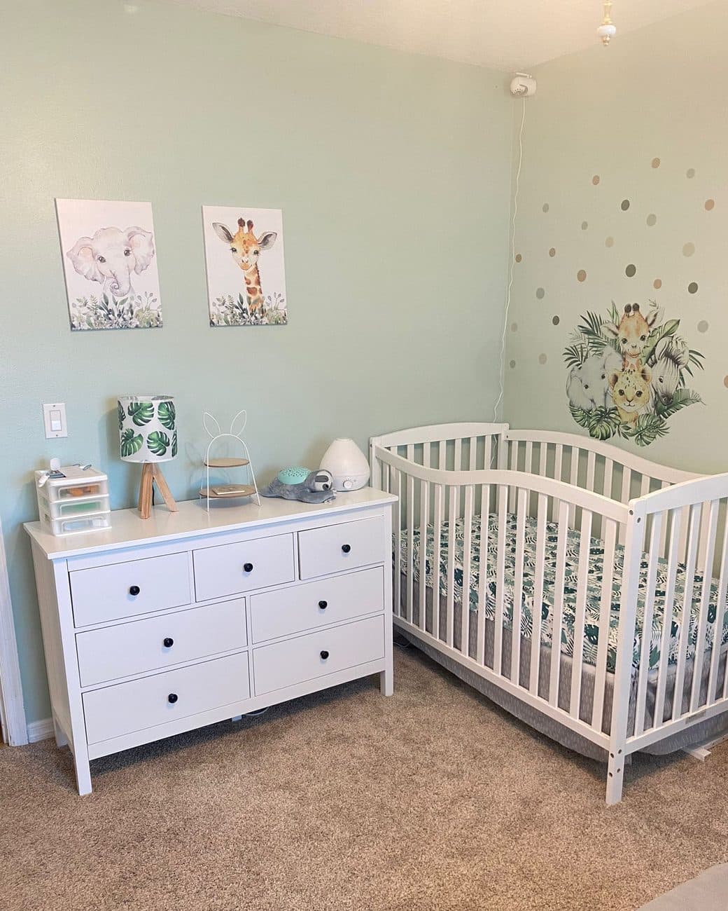 A finished nursery with a white dresser, a white crib and soft green walls, the dresser top holding only a lamp and a few small things.