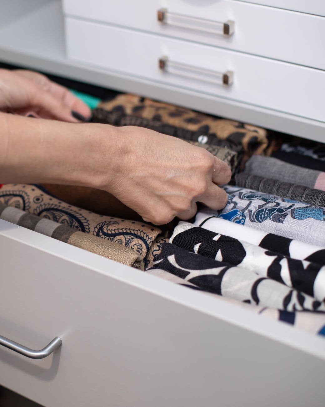 Hands folding a scarf into a wide white drawer where every piece already sits in its own row.