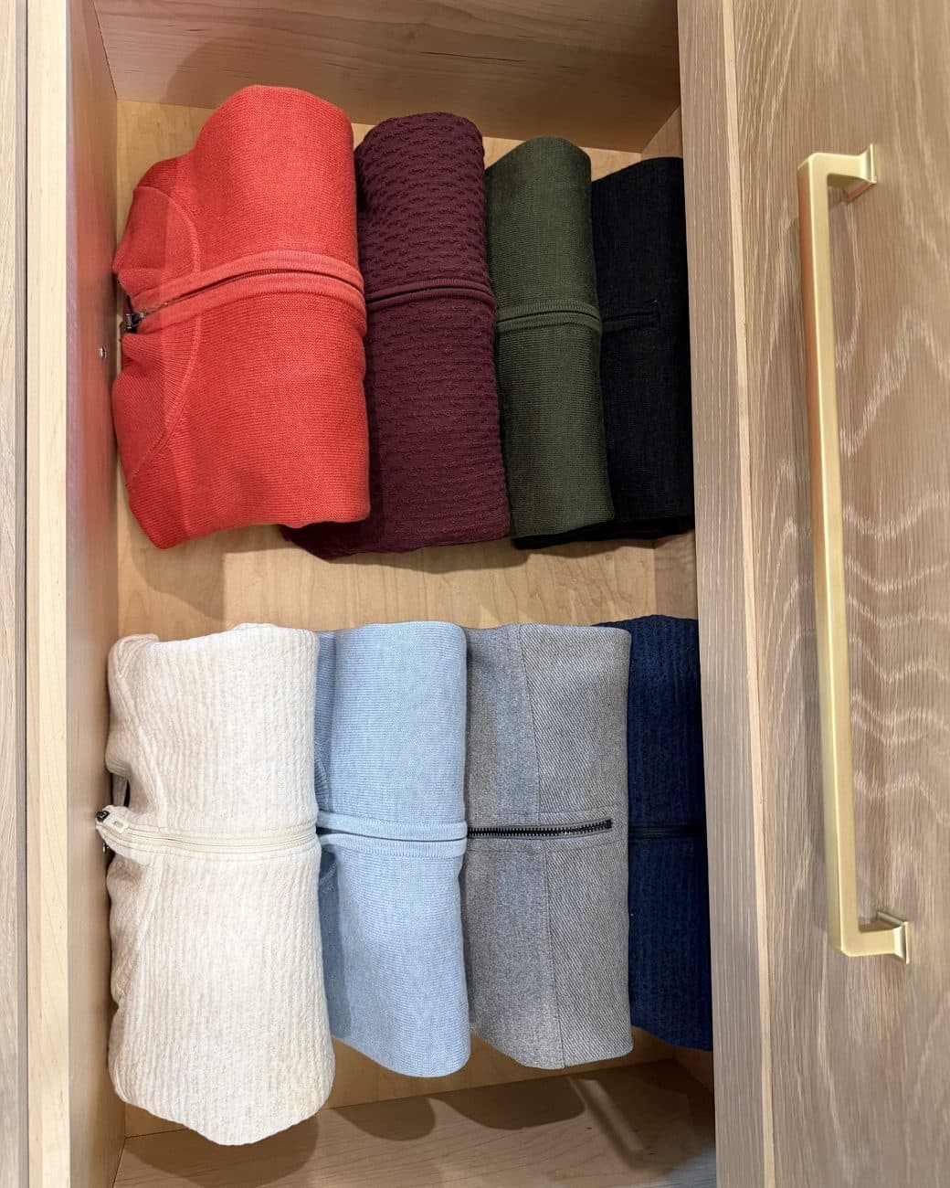 An open oak drawer with a brass handle holds eight folded sweaters, standing upright in two calm rows of color.