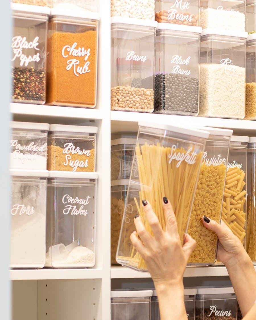 Hands slide a jar of spaghetti onto a pantry shelf lined with clear containers, each labeled by hand.